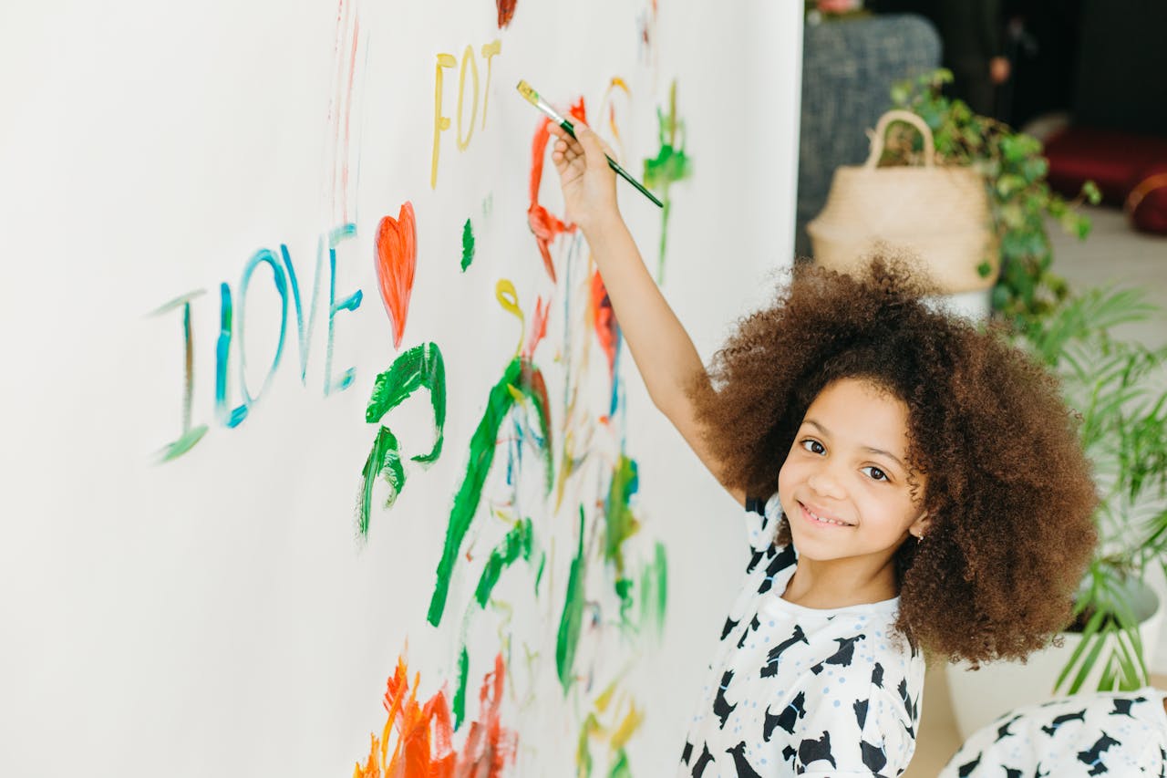Smiling girl with curly hair painting colorful words on a wall indoors.